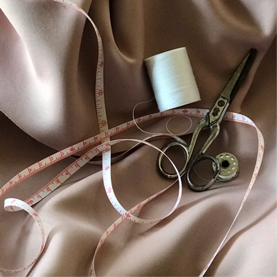 A pair of scissors, measuring tape, and two spools of white thread laid on top of a shiny sheet of fabric.